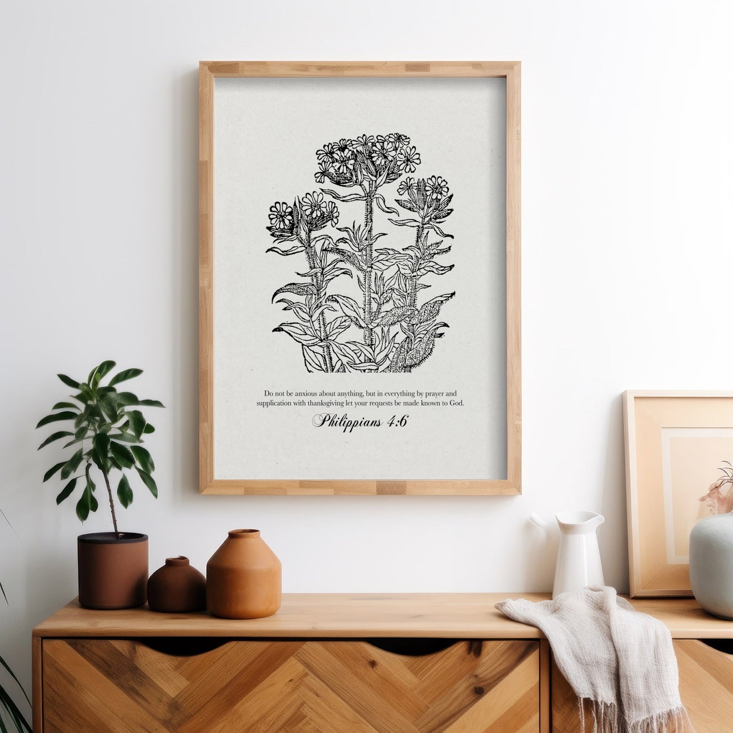 Philippians 4:6 botanical print with black floral illustrations, framed in wood and styled on a wooden dresser with terracotta decor and a small potted plant.