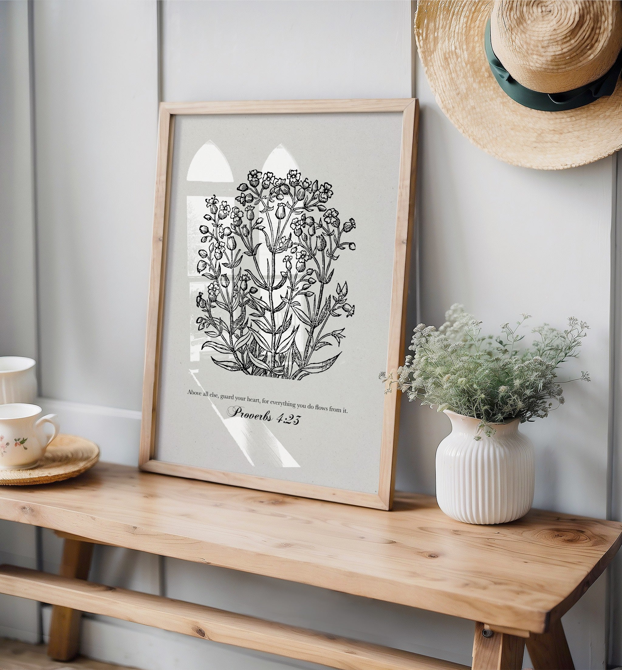 Black floral botanical art print featuring Proverbs 4:23, leaning on a rustic wooden surface with soft greenery and decor elements like a white vase and straw hat.