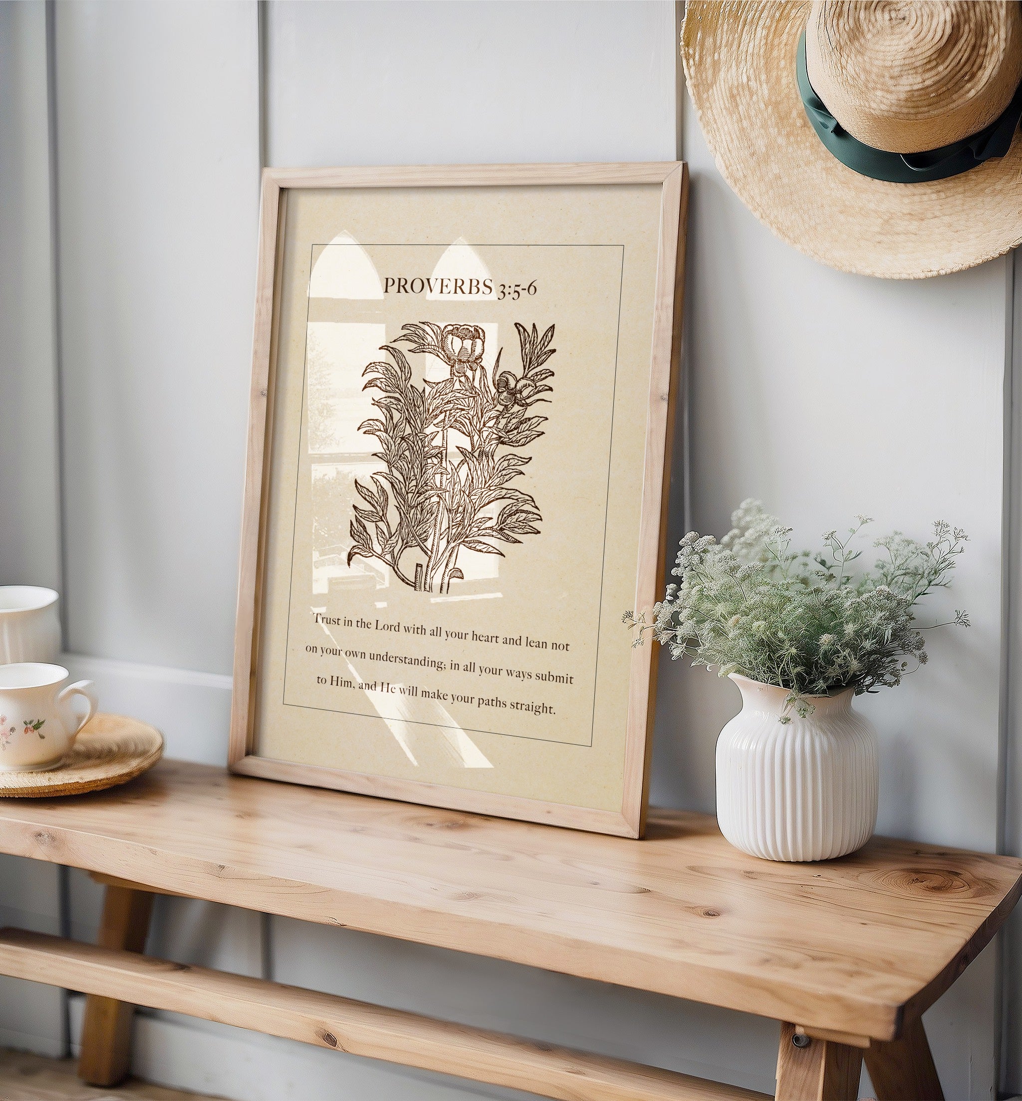 Vintage floral art print with Proverbs 3:5-6, leaning on a wooden surface alongside delicate decor like a white vase with greenery and a straw hat hanging nearby.