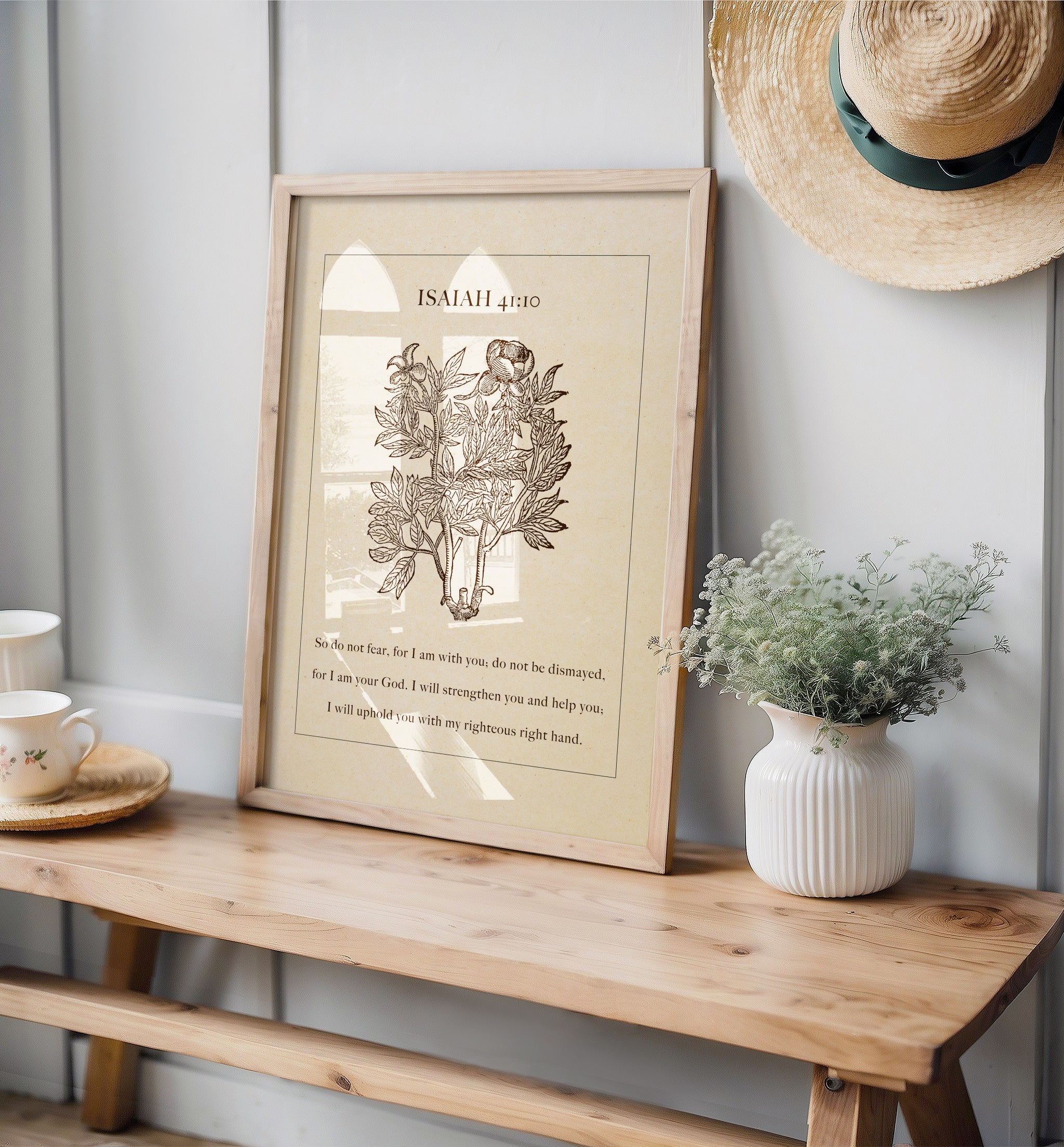 Isaiah 41:10 botanical art print leaning on a wooden bench, styled with a white vase of delicate greenery, vintage teacups, and a straw hat hanging on a light-colored wall, creating a serene and rustic setting.