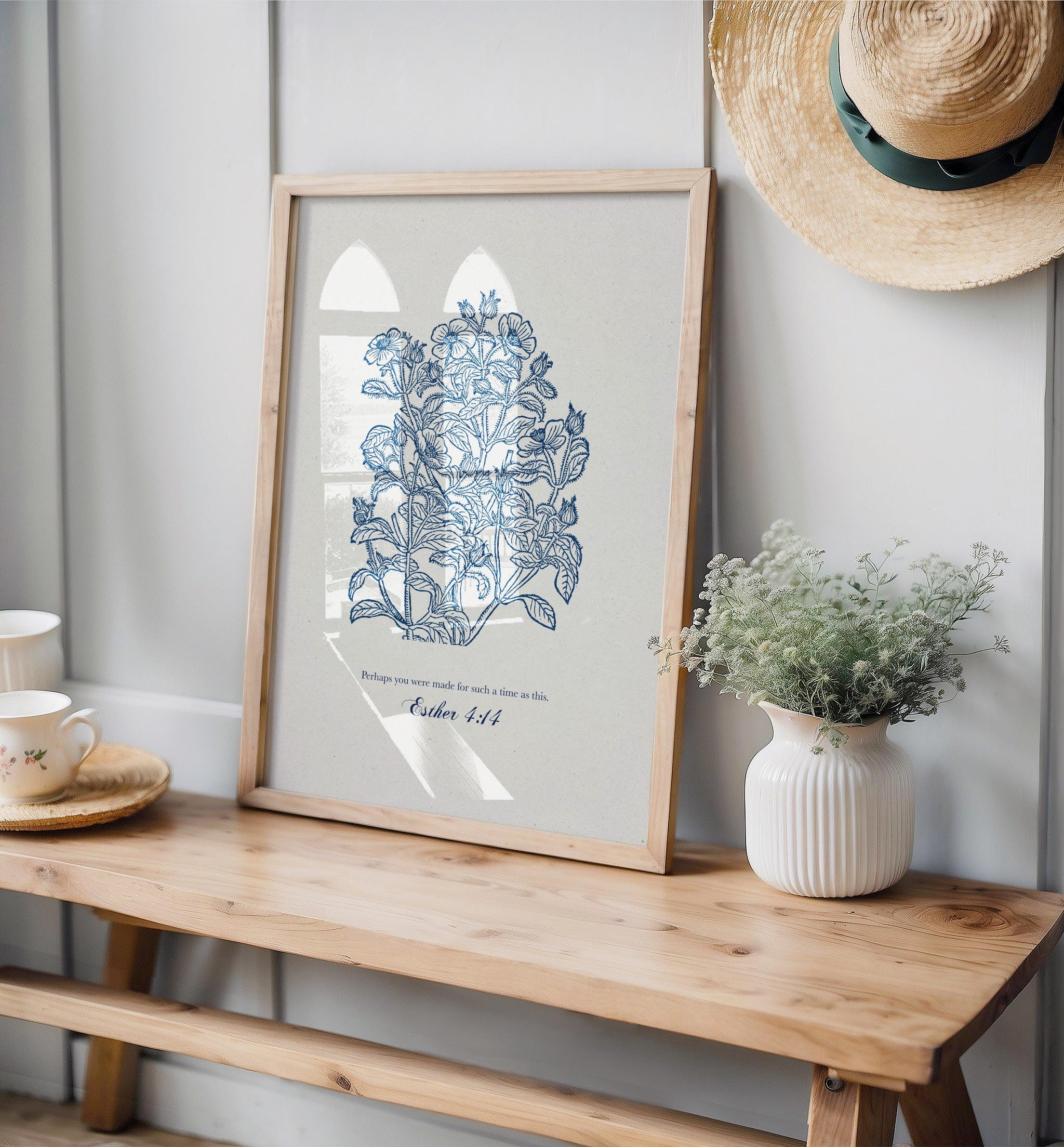 Framed Christian botanical print on a wooden bench with a vase of flowers and a hat on a gray wall.
