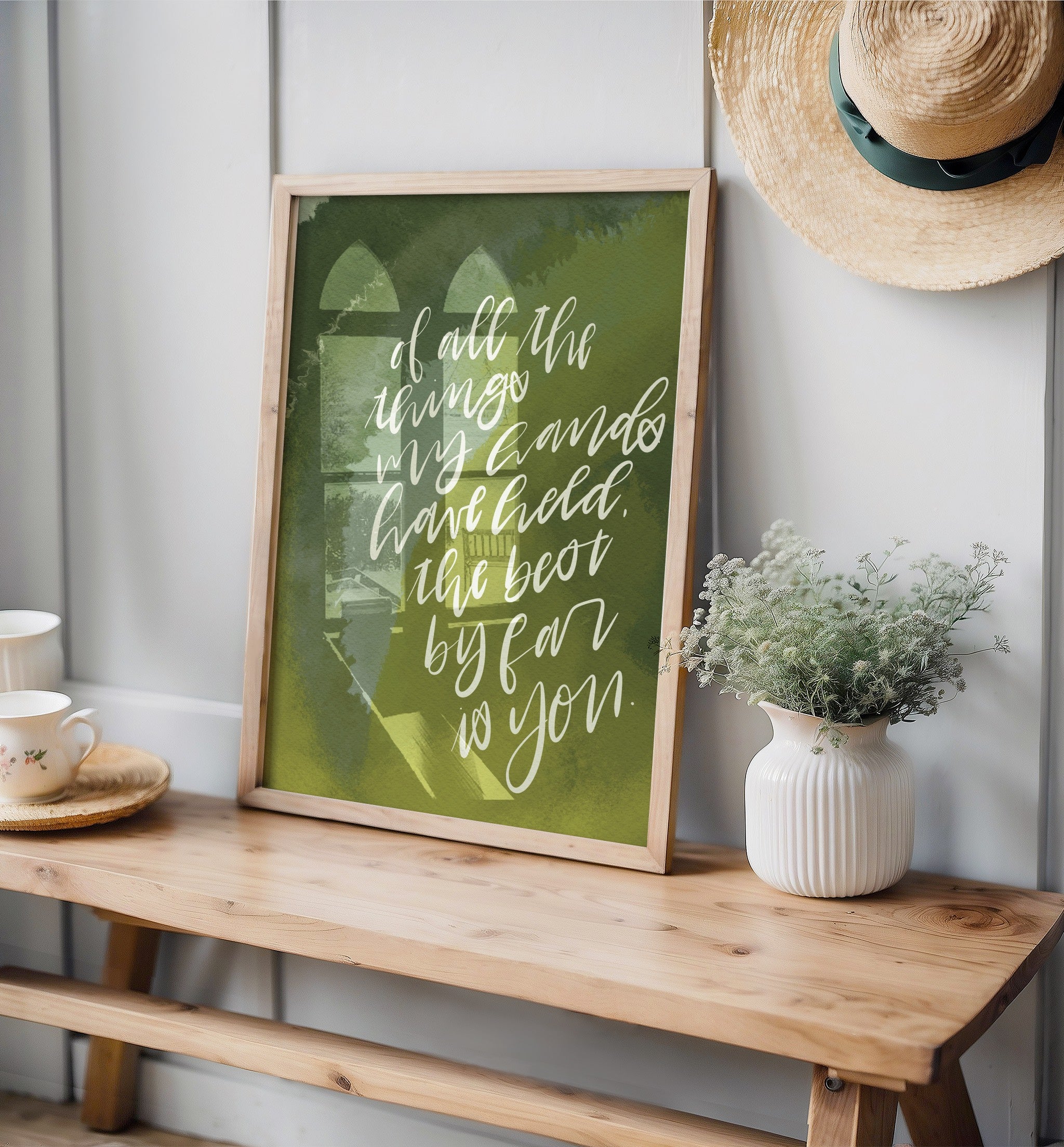 Framed artwork on a wooden bench with a vase of flowers and a hat in the background.