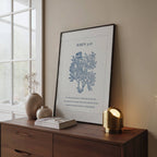 Framed artwork featuring John 3:16 Scripture and blue botanical on a wooden surface next to a window.