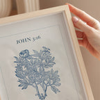 Framed print with 'JOHN 3:16' and a botanical illustration, held by a hand.