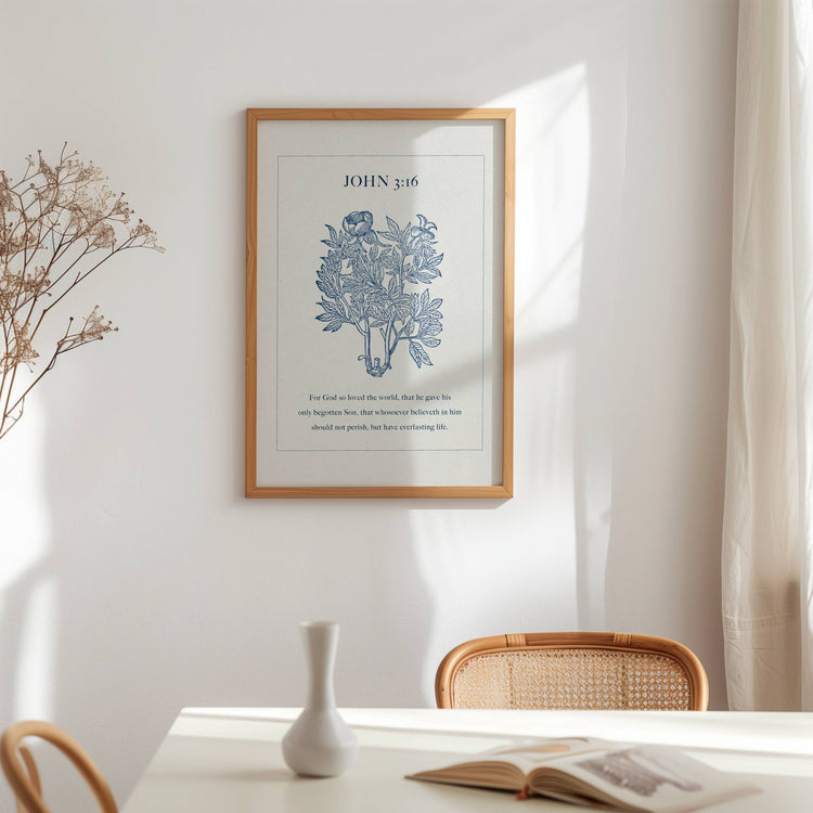 Framed artwork featuring John 3:16 Scripture and blue botanical on a wall above a table with a vase and open book.