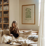 Woman using a laptop on a bed in a cozy room with a bookshelf and framed picture featuring John Muir Forest Wilderness quote and beautiful floral art by Reflourish Collective.