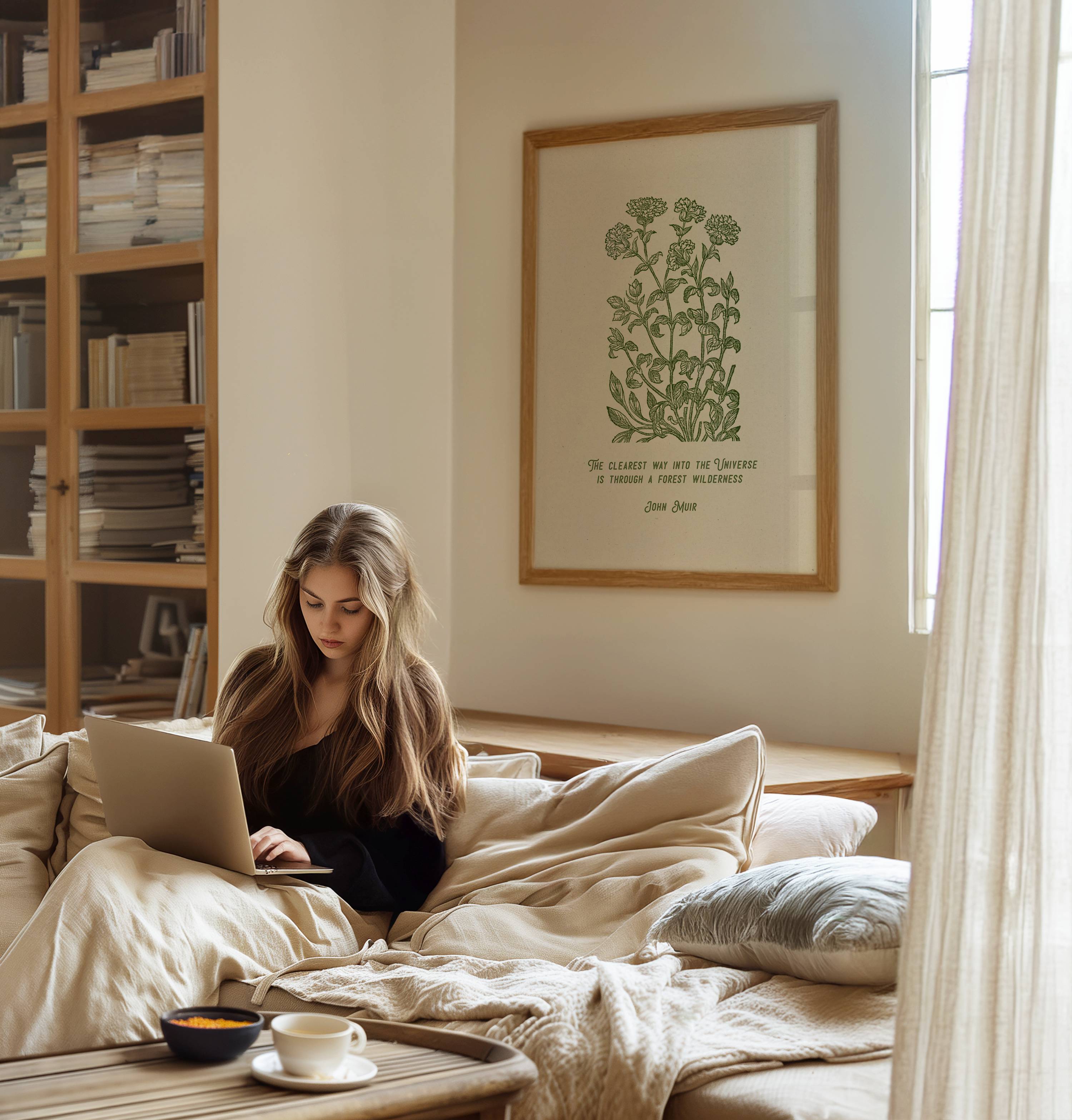 Woman using a laptop on a bed in a cozy room with a bookshelf and framed picture featuring John Muir Forest Wilderness quote and beautiful floral art by Reflourish Collective.