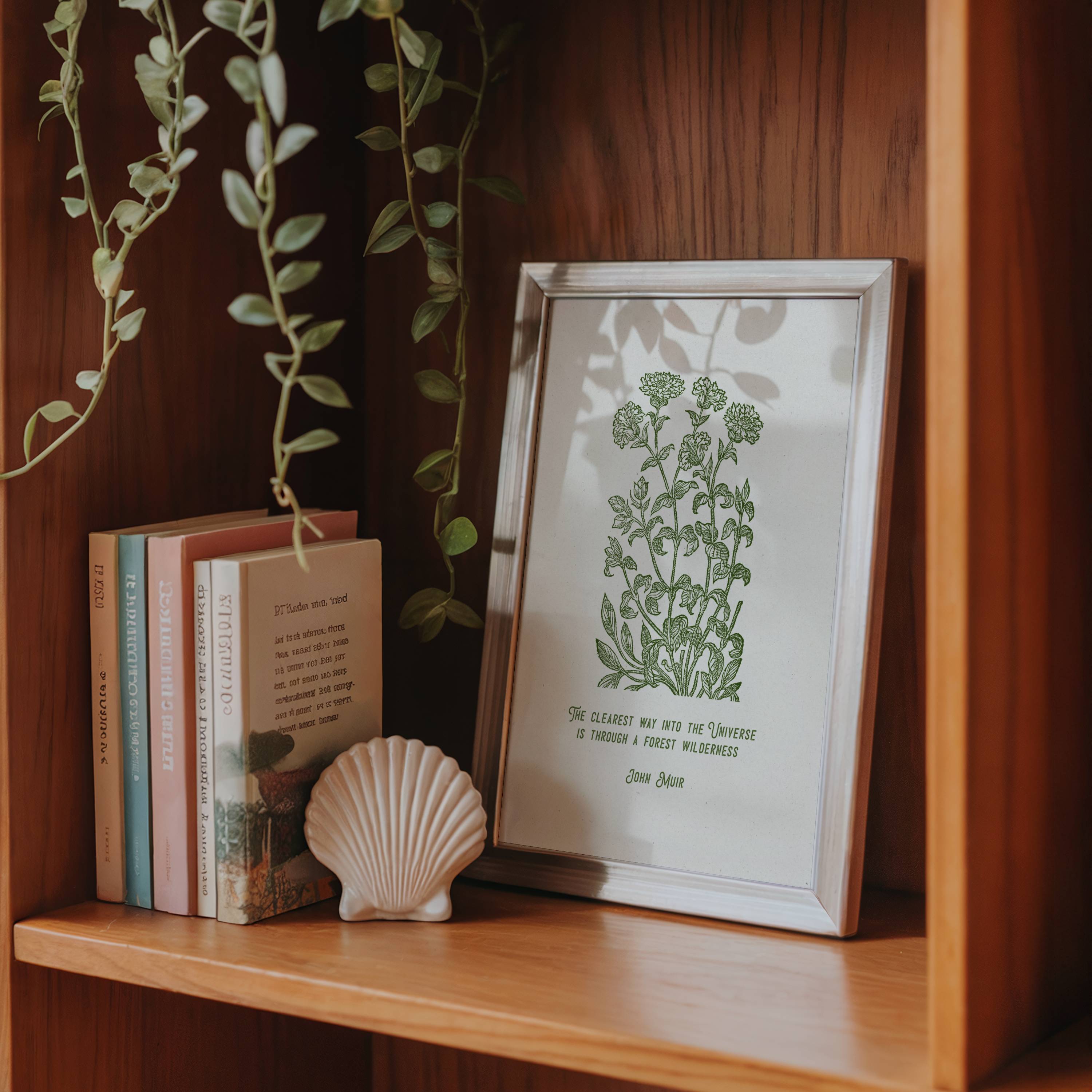 Framed print of green plants and John Muir quote shown on a wooden shelf with books and a shell.