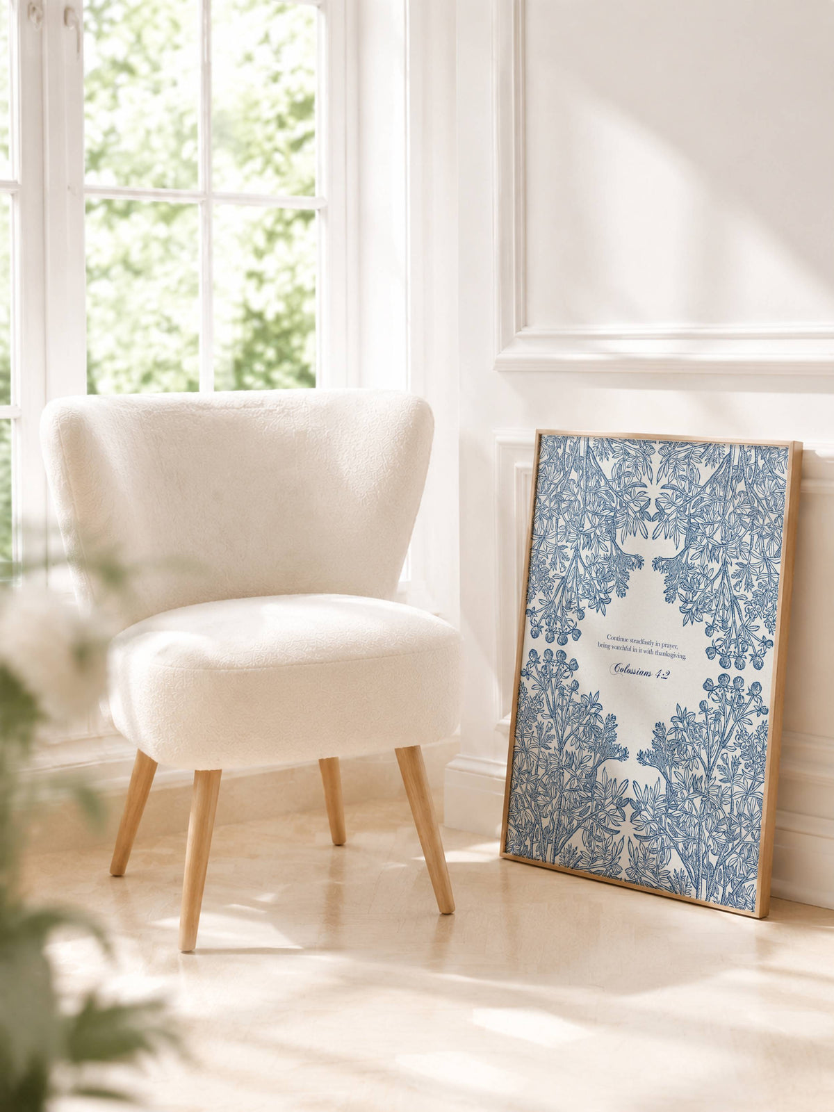 A framed blue and white botanical pattern art print leans against a white wall in a bright, sunlit room next to a cream-colored armchair.