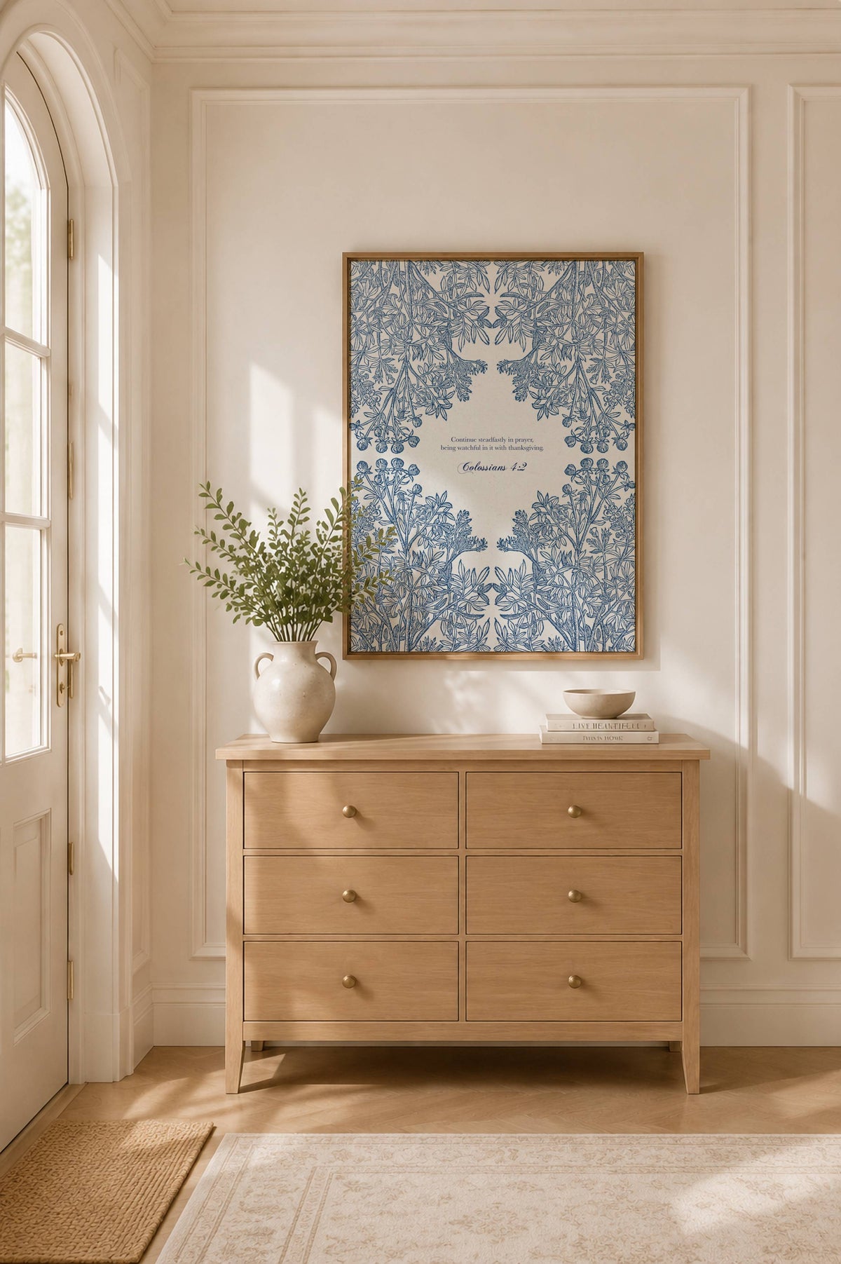 A framed art print featuring intricate blue floral patterns and Colossians 4:2 hangs on a cream paneled wall above a light wood dresser in a sunlit room.