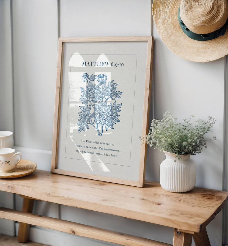 Framed artwork with Bible verse Matthew 6:9-10 on a wooden bench with a vase of flowers and a hat on a wall.