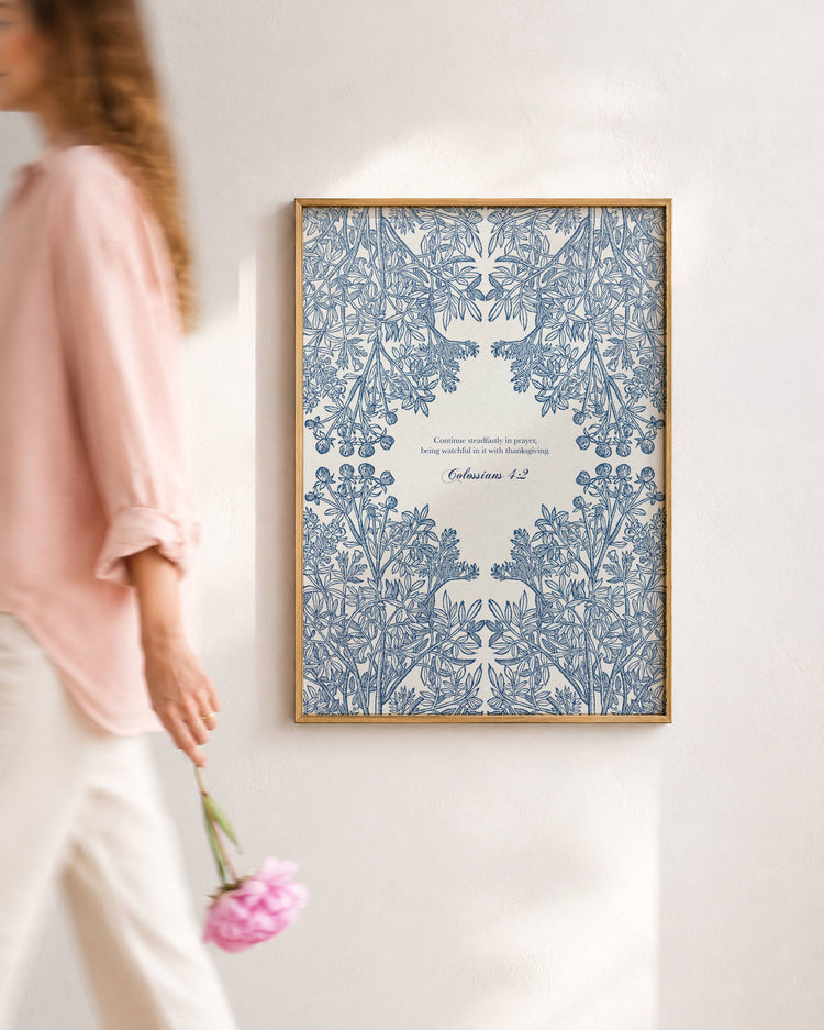 A framed art print featuring intricate blue floral patterns and Colossians 4:2, hung on a white wall. A woman in a pink top and white pants walks past on the left holding a pink flower.